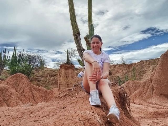 PauletteLoveX sitzt auf roten, rissigen Wüstensteinen, trägt weiße Turnschuhe, ein graues T-Shirt und weiße Shorts und lächelt. Im Hintergrund sind Kakteen und ein hoher, grüner Saguaro-Kaktus zu sehen..
