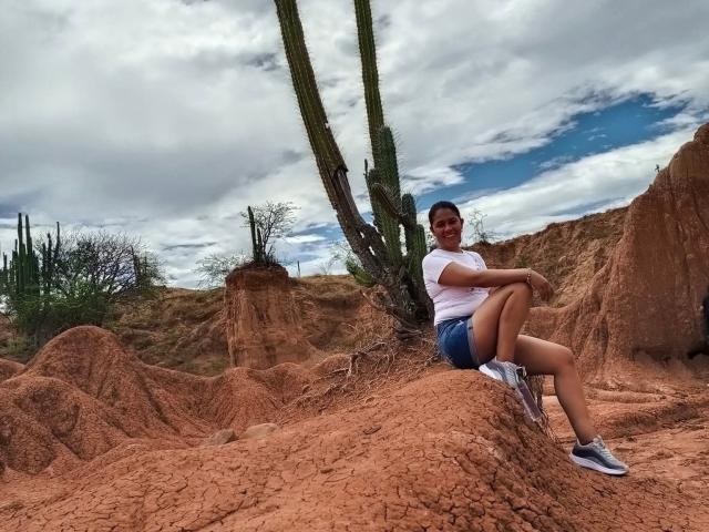 PauletteLoveX, sitzend auf rotem Felsen, trägt ein weißes T-Shirt, blaue Shorts und graue Turnschuhe, die Beine sind gekreuzt, sie lächelt, im Hintergrund sind Kakteen zu sehen, der Himmel ist bewölkt, die Arme liegen auf den Knien..