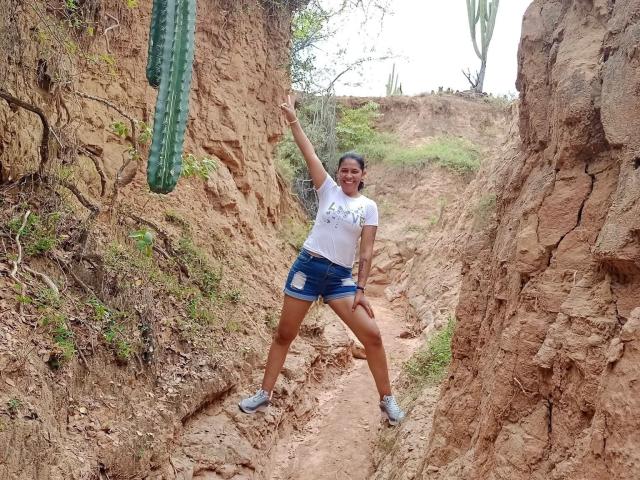 PauletteLoveX, parada em um desfiladeiro estreito de terra, vestindo uma camiseta branca e shorts azuis, com um braço levantado, sorrindo e apontando para cima, exibindo suas nádegas bronzeadas e tonificadas..