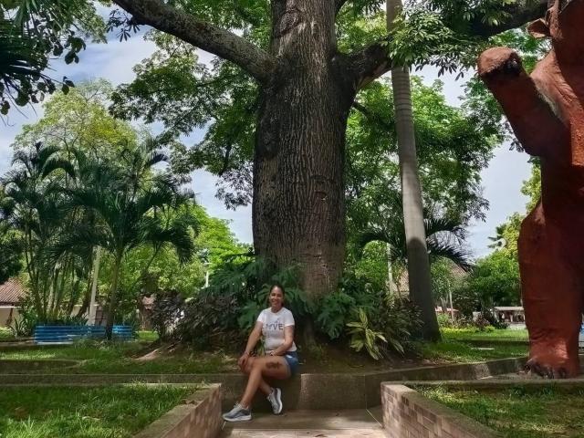 PauletteLoveX sits against a giant tree with her ass on a concrete ledge, wearing a white shirt and blue shorts, with greenery and a red statue in the background.