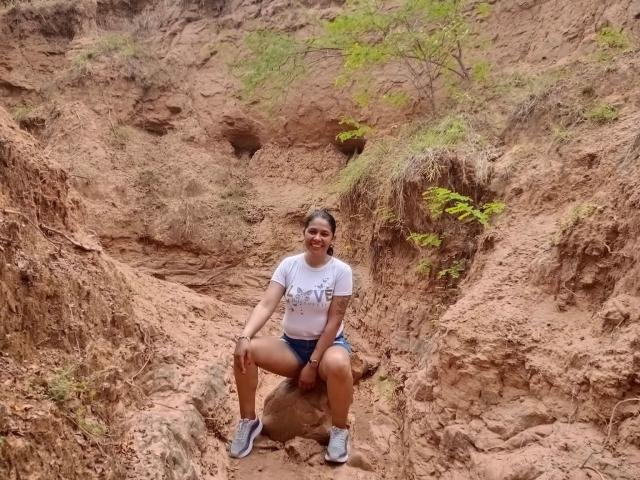 PauletteLoveX sitzt auf einem Felsen aus Erde, trägt ein weißes T-Shirt, blaue Shorts und graue Turnschuhe, mit gespreizten Beinen, und lächelt in die Kamera, in einer rauen, braunen, felsigen Gegend..