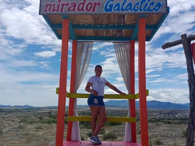 PauletteLoveX stands with a small ass and white t-shirt, holding a yellow rail, wearing blue shorts, and sneakers, under a colorful wooden structure with orange columns.