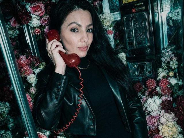 JakyKidman, avec des cheveux foncés, une veste en cuir noir, tenant un téléphone rouge à fil, portant un haut noir, entourée de fleurs, souriant légèrement, regardant la caméra..