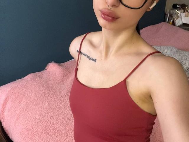 GlitchDoll with fair skin, small tits, and a nose ring, wearing a red tank top, has a tattoo on her chest, and is sitting on a pink textured surface.
