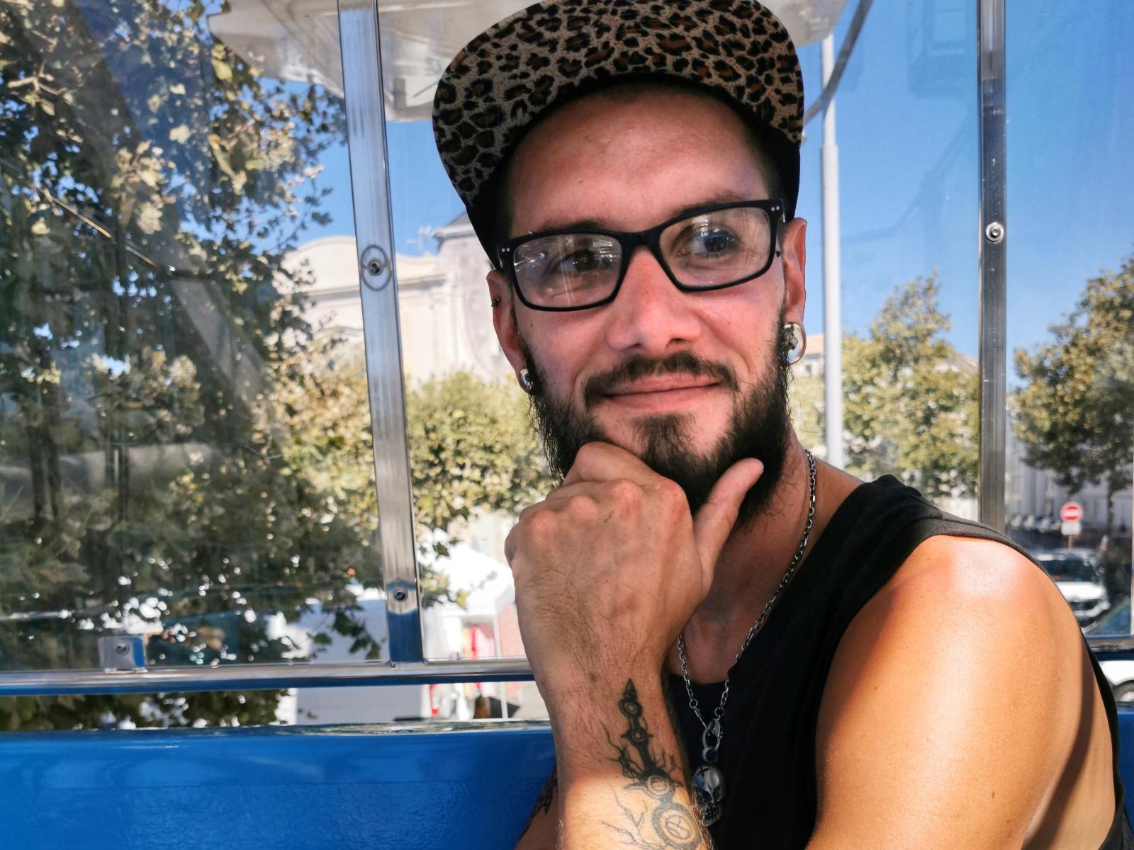 XBobbyNightX con un sombrero de leopardo, gafas negras, barba, tatuajes y una camiseta negra sin mangas, sonriendo, con la mano en la barbilla, fondo azul, soleado, y usando un collar de cadena..