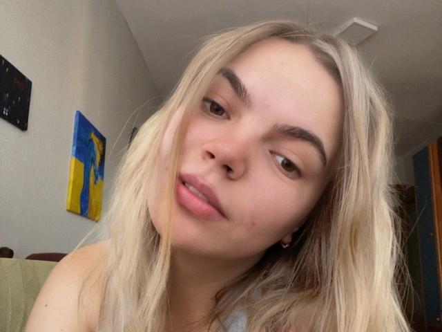 SoniyaS, com cabelo loiro, lábios levemente entreabertos e olhos castanhos, olhando para a câmera com uma expressão relaxada. Ela tem um nariz pequeno e está vestindo uma blusa cinza..