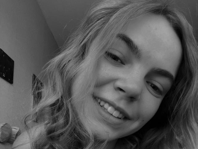 Black and white selfie of SoniyaS with wavy, blonde hair, smiling with visible teeth, slightly squinted eyes, and raised eyebrows, showing a relaxed and playful expression.