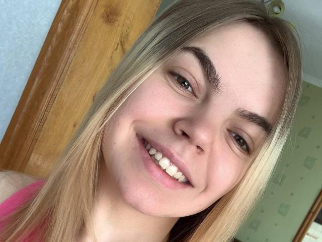 SoniyaS has blonde hair, fair skin, and is smiling with her pink lips and white teeth. She's wearing a pink top, showing her cute face and brown eyes.