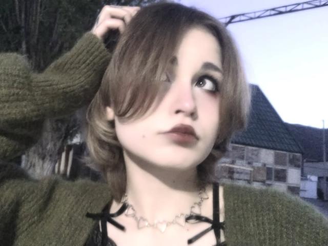 Emely Dilsaver tem cabelo castanho curto, um suéter verde, uma blusa preta com tiras, um colar de prata e uma pequena marca na pele no queixo, olhando para o lado com a mão no cabelo..