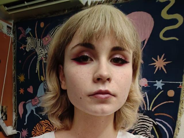 EmelyDilsaver has blonde hair, pink eyeliner, freckles, and a small mole on her chin, with a neutral expression, wearing a white shirt, in a colorful, zebra-patterned background.