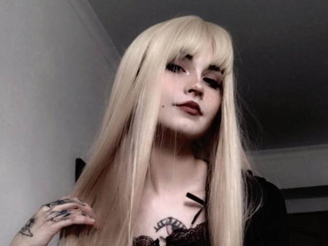 EmelyDilsaver has long blonde hair, dark eyeliner, and a small nose piercing. She's wearing a black lace top, showing tattoos on her chest and hand.