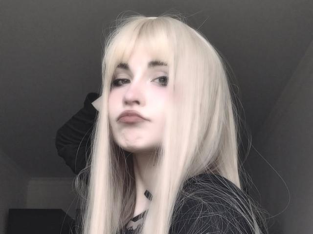 EmelyDilsaver with pale blonde hair, full lips, and a slight smirk, wearing a black top, has a small black tattoo near her neck, in a soft, natural light.