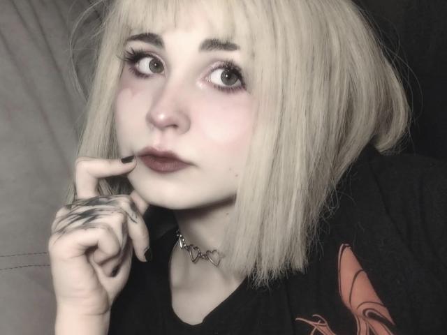 EmelyDilsaver has pale skin, blonde bob, and is looking up with wide eyes, touching her lip with a tattooed hand. She wears dark lipstick and a choker.