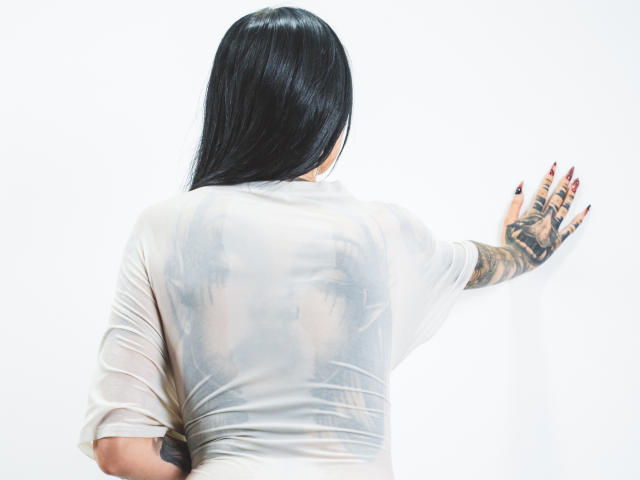 LeilaWiliam, con i capelli neri, di spalle alla fotocamera, indossa una camicia trasparente bagnata che mostra tatuaggi sul braccio destro, sfondo bianco, una mano appoggiata al muro, unghie dipinte di rosso..