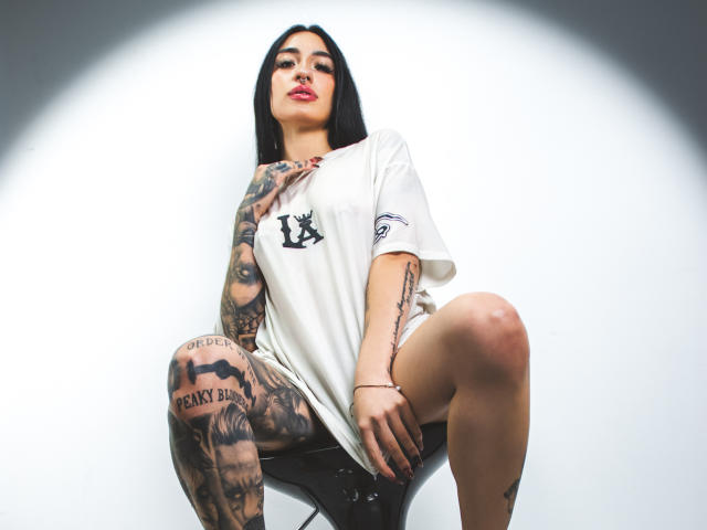 LeilaWiliam with tattoos, wearing a white tee, sitting with legs apart, black chair, black hair, red lips, looking down, right arm tattooed, left thigh tattooed, white background.