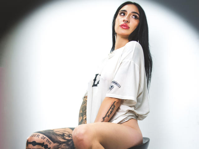 LeilaWiliam, con lunghi capelli neri, rossetto rosso, maglietta bianca, tatuaggi sul braccio e sulla coscia, seduta su uno sgabello nero, che guarda verso l'alto, mostrando la vulva scoperta, gambe tatuate, sfondo bianco..