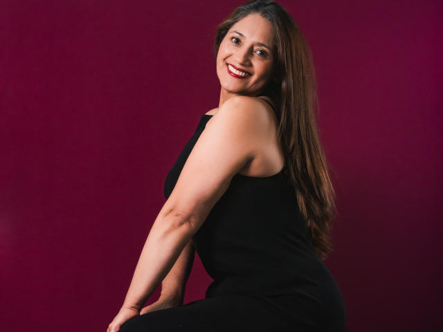 MarciaMuller, com cabelos castanhos longos, sorrindo, vestindo um vestido preto sem mangas, mostrando o bumbum e os seios, sentada em frente a um fundo vermelho..