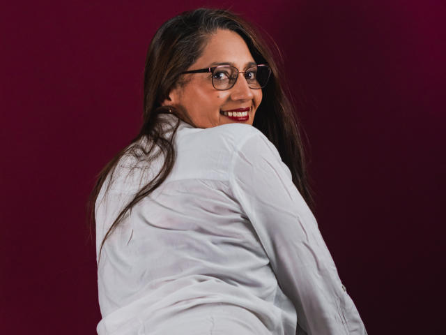 MarciaMuller met lang bruin haar, een bril, een wit shirt, die over haar schouder kijkt en terugkijkt, met een rode achtergrond, waarbij haar achterwerk zichtbaar is, gekleed in een shirt, en die er schattig en aantrekkelijk uitziet..