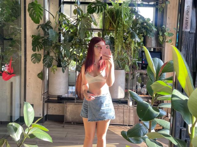 AlexXas, avec ses cheveux roux, ses lunettes, son soutien-gorge blanc et sa jupe en jean, prend un selfie devant un miroir, montrant ses seins et ses fesses fines et jolies, entourée de plantes vertes, et tenant un téléphone rose..