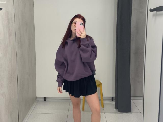 AlexXas, avec des cheveux roux, des lunettes, un sweat à capuche violet, une jupe noire, tenant un téléphone dans un miroir, montrant ses fesses, dans une pièce minimaliste, debout à côté d'un tabouret jaune..