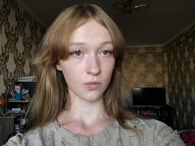 Kimbra Renuart, avec sa peau pâle, ses taches de rousseur et ses yeux verts, portant un haut beige, a de longs cheveux blonds et une expression légèrement boudeuse, dans une pièce peu éclairée..
