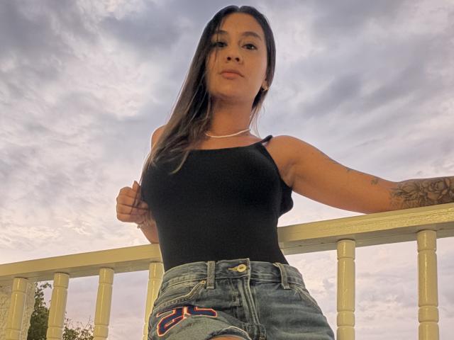 ThaliaAres with long brown hair, black tank top, tattooed arm, wearing denim shorts, standing against a white railing, looking down with a serious expression, cloudy sky in background.