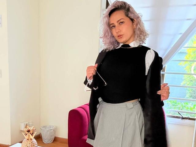 Elena Saenz, con cabello rizado rosa, suéter negro, camisa blanca, falda gris y abrigo negro, sosteniendo gafas, de pie frente a una silla rosa, suelo de madera y una ventana..