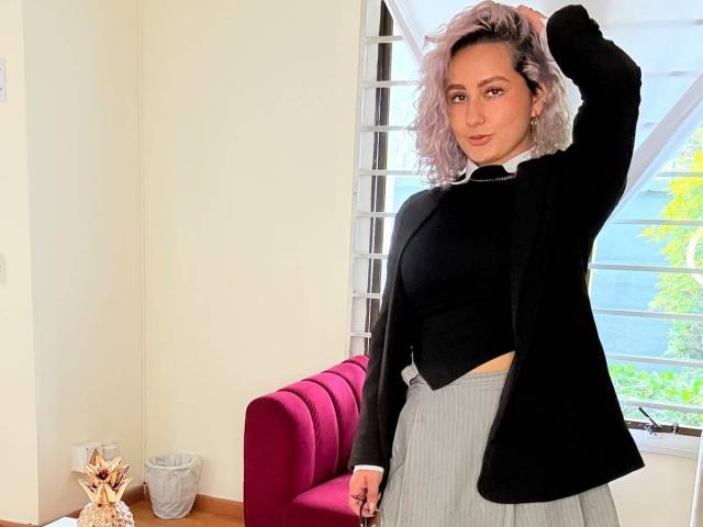 Elena Saenz, com cabelo grisalho rosado, blusa preta, camisa branca, saia cinza e jaqueta preta, em pé, com a mão no cabelo, em frente a uma cadeira rosa, janela, piso de madeira e parede branca..