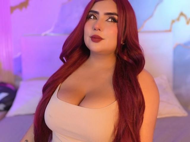 AriiStonee with long red hair, large tits in a beige tank top, looking to the side with a slight smile, in a softly lit bedroom.