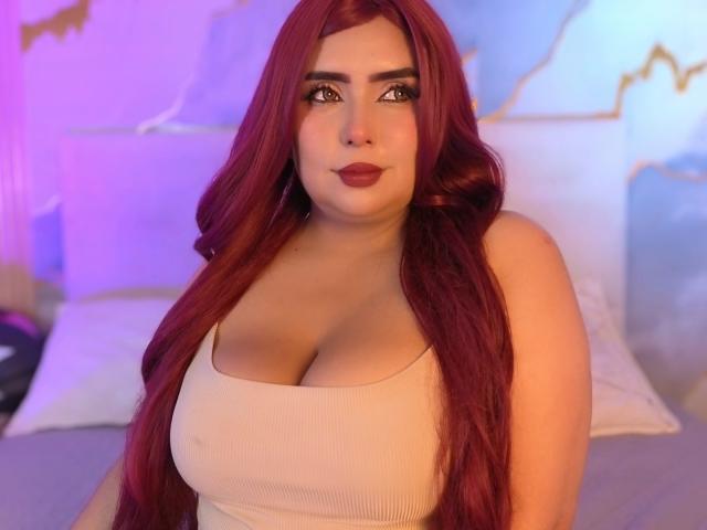 AriiStonee with long red hair, big tits, and a cute face wearing a beige tank top, sitting on a bed with a soft purple and blue light.