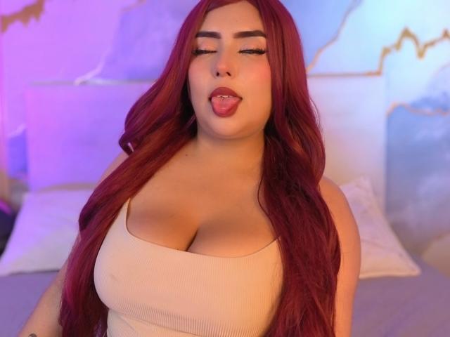 AriiStonee with long red hair, tongue out, wearing a beige ribbed tank top, showing big tits, pink and blue lights, and white pillows in the background.