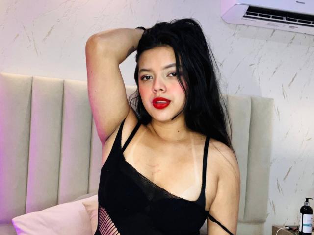 ViolettaRouse with long black hair, red lipstick, and black lingerie, showing her tits, with one arm raised, sitting on a bed, air conditioner above her.