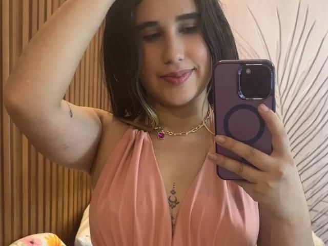 KatieDesirX, con i capelli scuri, un vestito rosa, una collana d'oro e un tatuaggio sotto il seno, sta scattando un selfie allo specchio con un telefono viola, sorridendo con un braccio alzato..