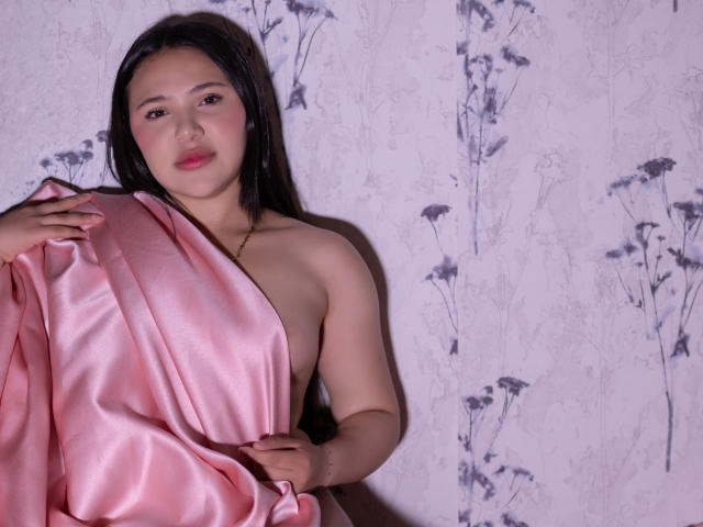 NicolDanniels with pink satin covering her tits, holding it with one hand, black hair, light skin, pink lipstick, standing against a floral-patterned wall, looking at camera.