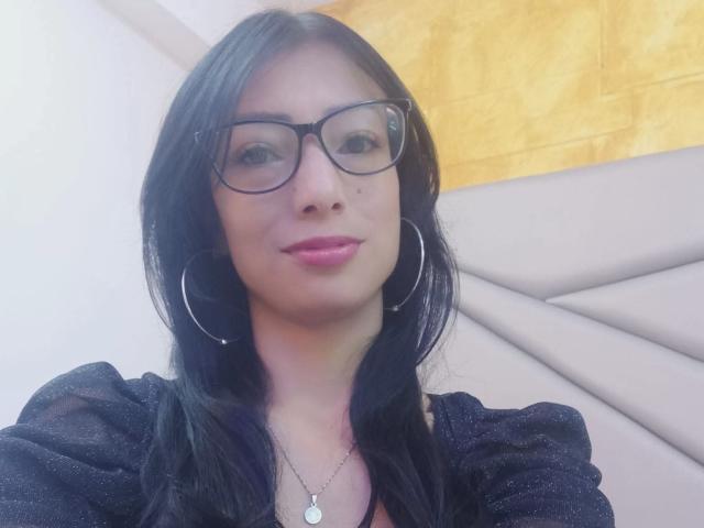 AlejandraCherry has black hair, glasses, and pink lipstick, wearing a black top with puffy sleeves, and large silver hoop earrings, smiling softly.