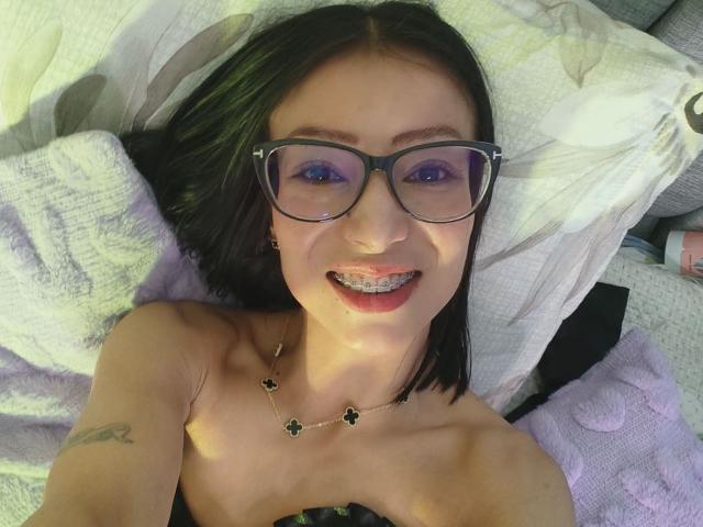 AlejandraCherry, avec des lunettes noires, souriant, montrant ses dents avec du dentifrice blanc, allongée sur un oreiller, portant un collier, a un tatouage sur son bras droit, et une bandoulière noire..