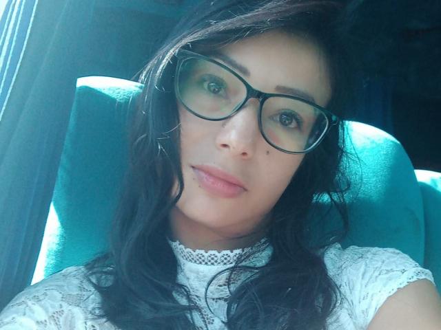 AlejandraCherry, avec des lunettes noires, des cheveux noirs ondulés, une robe blanche en dentelle, des lèvres roses et une petite beauté sur sa joue gauche, assise sur une chaise turquoise..