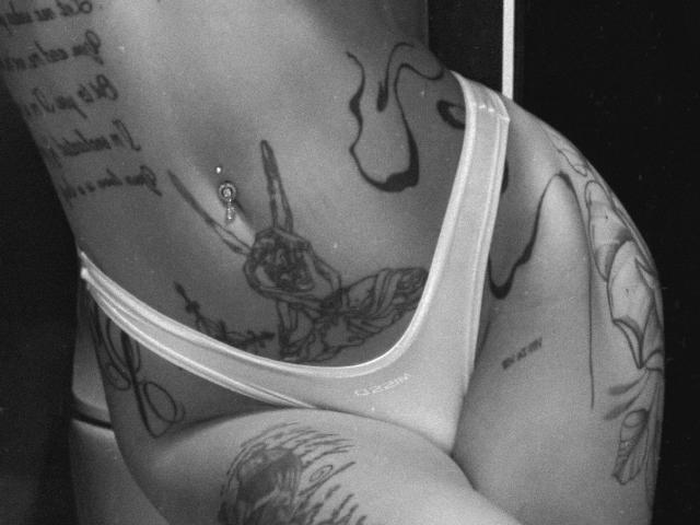 Black-and-white photo of SensualMila's tattooed lower back and buttocks in sheer white panties. She has a navel piercing, tattoos including a hand and text, and a curvy ass.