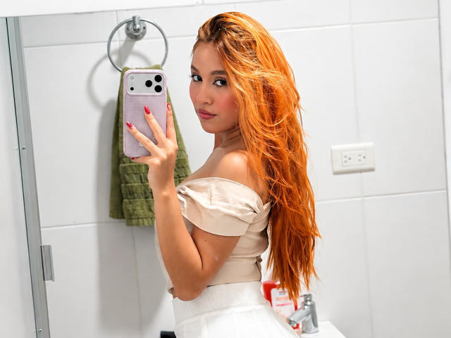 EmmaWat with long orange hair, off-shoulder beige top, white skirt, taking a mirror selfie with pink iPhone, red nails, green towel, and white bathroom background.