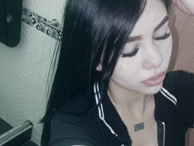 NyraVale, com cabelo preto, olhos fechados e cílios longos, vestindo uma jaqueta preta com zíper, um piercing no nariz e um colar de prata, mostrando uma tatuagem abaixo do queixo..