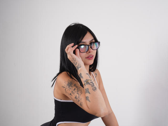 NyraVale with black hair, glasses, and tattoos on arms, touching her face, wearing a black sports bra, showing her tits. White background.