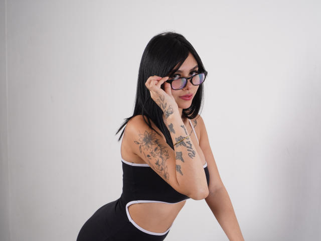 NyraVale with black hair, glasses, and tattoos, wearing a black sports bra and matching shorts, adjusting her glasses, showing her tits and a cutout in her shorts.
