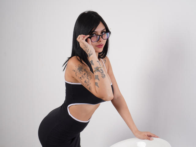 NyraVale, con cabello negro, gafas y tatuajes, vestida con un vestido negro con aberturas, apoyada en una superficie blanca, mostrando su trasero curvilíneo y sus pequeños senos..