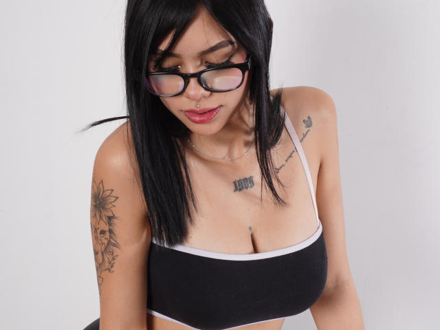 NyraVale, avec ses cheveux noirs, ses lunettes et son anneau de nez, porte un débardeur de sport noir, dévoilant ses seins, des tatouages sur le bras gauche et la poitrine, ainsi qu'un petit tatouage sur l'épaule droite..