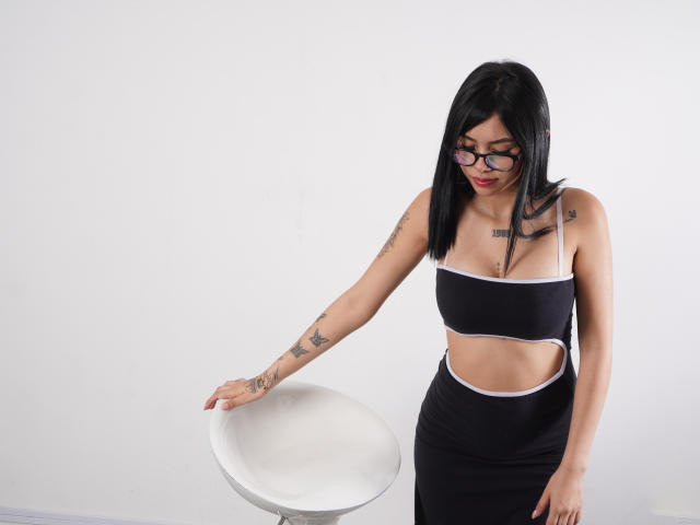 NyraVale, con cabello negro y gafas, vestida con un vestido negro con aberturas, tocando una mesa circular blanca, con tatuajes en los brazos y lápiz labial rojo, de pie en una habitación sencilla..