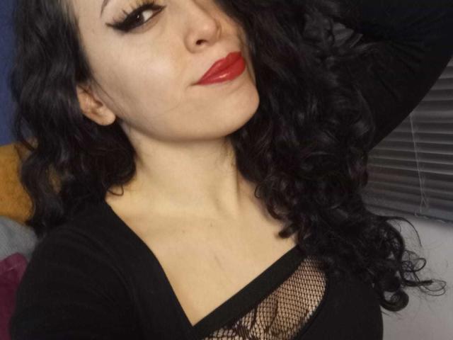 KendraSimons, con i capelli neri ricci, rossetto rosso e top in rete nera che mostra il seno, braccio alzato, che guarda la fotocamera, con trucco scuro per gli occhi, pelle chiara, in una posa leggermente inclinata..