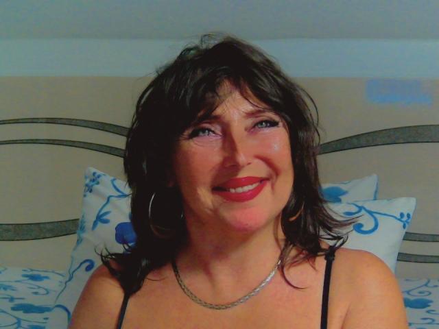 Ossenna tiene el cabello castaño oscuro y ondulado, lápiz labial rojo y un collar de plata, sonríe con ojos azules, lleva un top negro con tirantes y grandes aros..