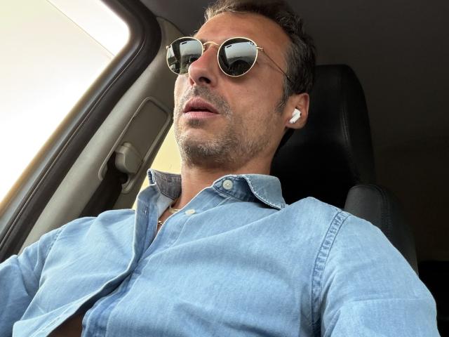 Morus with a light blue denim shirt, round sunglasses, and white earbuds, sitting in a car, looking forward with a slightly open mouth. His ass is not visible.