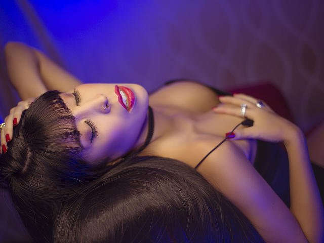 SeleneBrigth with her eyes closed, dark hair, red lipstick, black choker, and red nails, lying back, hands on her tits, wearing a black strap dress, in a blue-lit shot.