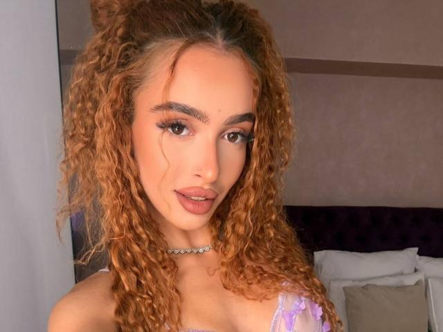 MarissaFex with curly brown hair, wearing a purple lace top, choker, and makeup, looking at the camera with a slight smile, showing her tits.